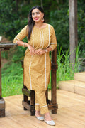 A woman standing on a wooden surface wearing a chestnut floral kurta set with straight hemline and side slits, round neckline, and 3/4 sleeves with scalloped lace details. She is also wearing straight-leg pants and white shoes.